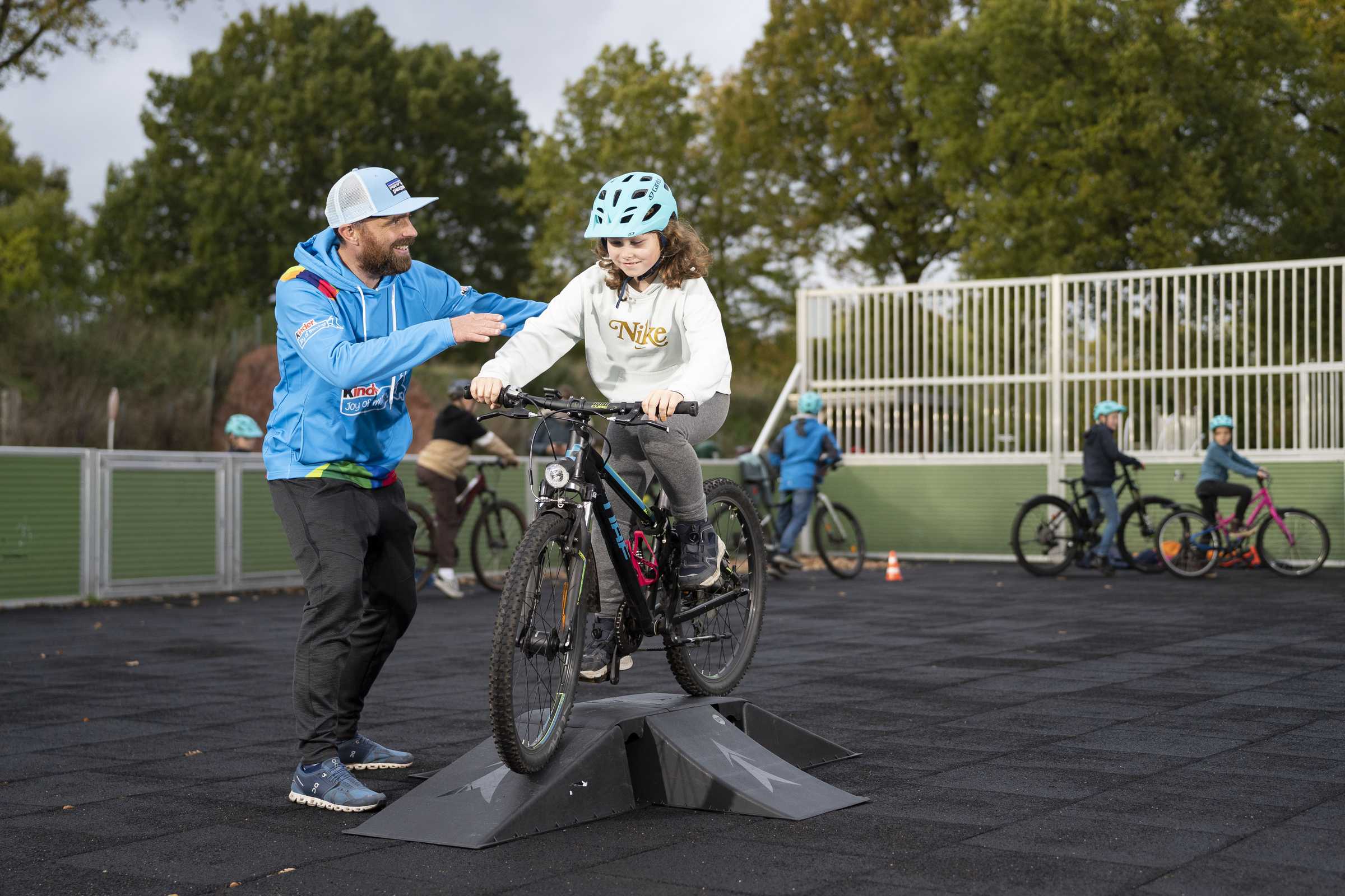 Ein Trainer begleitet ein Mädchen mit Fahrradhelm über eine Rampe, im Hintergrund weitere Kinder auf Rädern