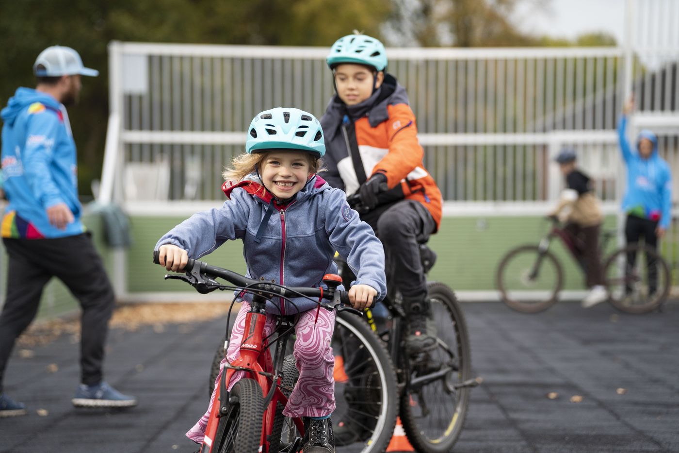 Ein Mädchen mit Fahrradhelm fährt lachend auf die Kamera zu, dahinter ein Junge auf dem Rad und zwei Trainer auf einem abgesperrten Platz